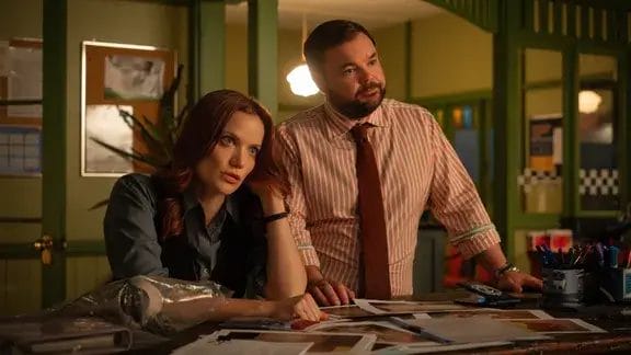 New Detective, New Mystery: Return to Paradise Brings Aussie Crime Drama Spin-off to the Death in Paradise Universe Premiering Soon on BBC One and iPlayer 19 Return to Paradise still which sees Anna Samson as Mackenzie Clarke sitting at a table with Lloyd Griffith as Detective Colin Cartwright standing beside her
