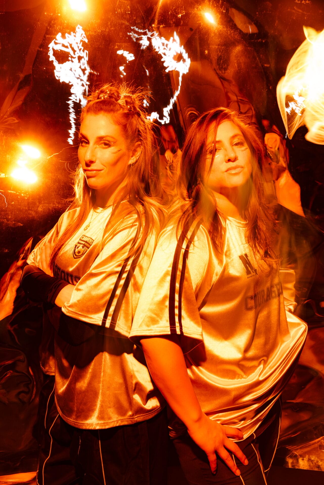 Full portrait image of duo Royal She posing wearing gold tops and illuminated in a golden glowing lights with a gold filter.
