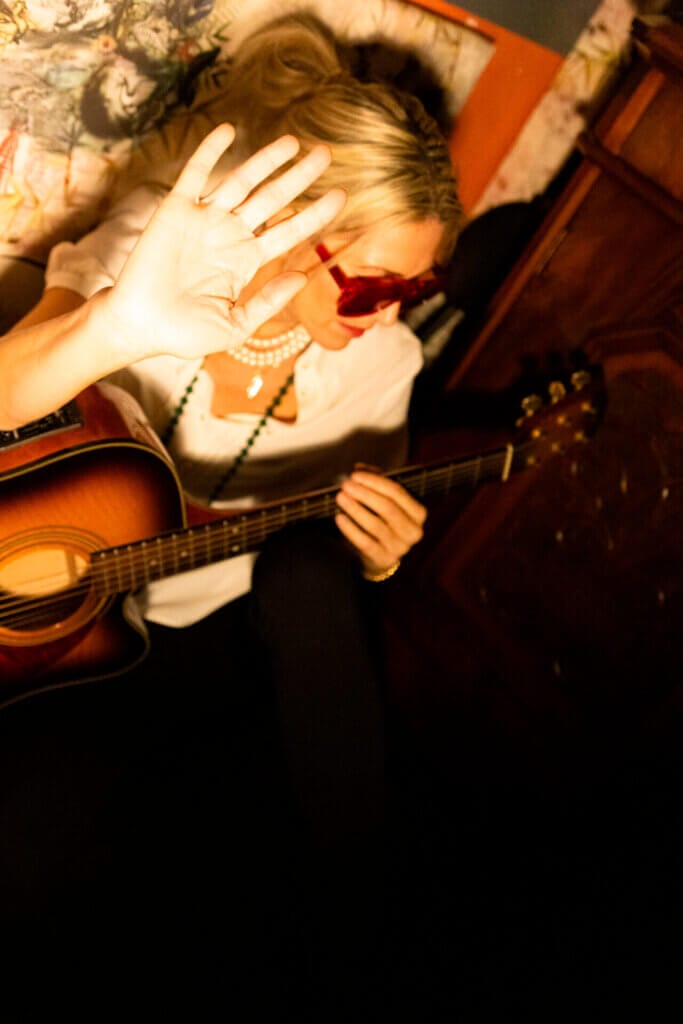Bird Revives "Falling Like Stars" to Mark 20 Years Since Her Debut Album With an Awesome 2024 Remaster 18 Bird sitting on the floor, turning her head away and holding up her hand to the camera, with her guitar across her lap.