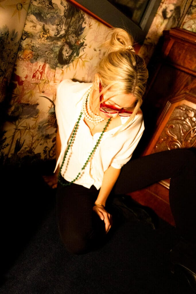 Bird sitting on the floor in a cream blouse and dark leggings, with red shades sitting against a Japanese art painting. She has her head pointed down and looks like she's about to get off the floor.