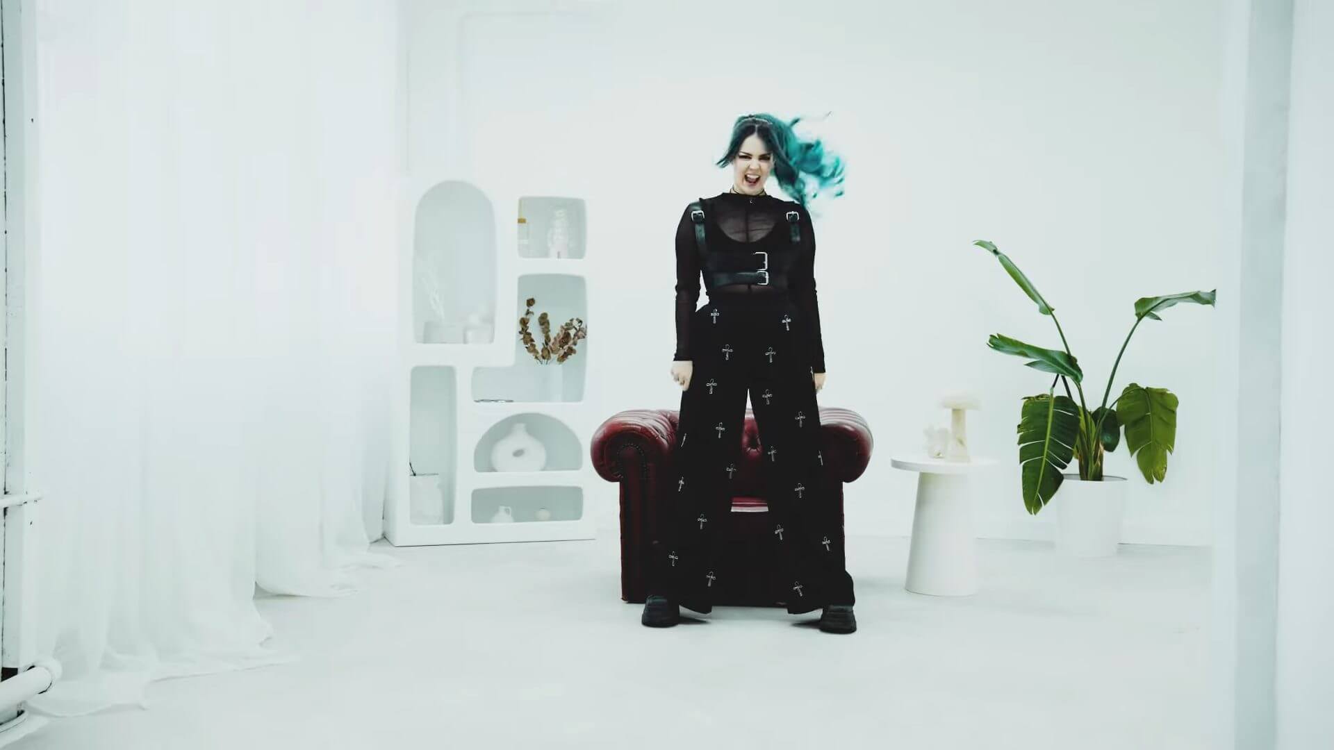 "Therapy": As December Falls Delivers a Hard-Hitting Anthem With Raw Relatable Energy and an Explosive New Music Video 20 Still from the "Therapy" music video which sees frontwoman, Bethany Hunter, standing in a white room with a brown leather chair behind her and a green plant to her left.