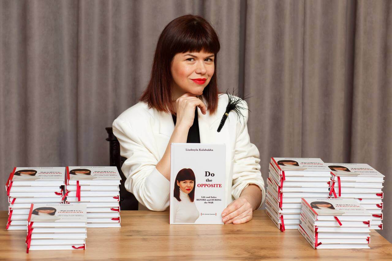 Liudmyla Kalabukha posing at a table, wearing a white jacket, with her right elbow on the table as she rests her chin on the right's fingers. On the table are six piles of her book, "Do the Opposite: Life and Sales Before and During the War", with one out in front of her.