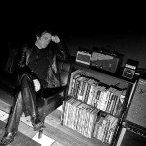 Black and white photo of Justin Climer who created the Mountain Cllimer project, sitting on a chair, dressed smartly, with a stereo to his left which has books and records on a shelf, underneath.