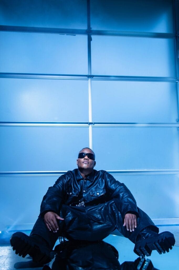 Sean Garrett Joins 2025 TRACE Awards & Summit as Panellist and Presenter 14 Sean Garrett, wearing black clothing paired with dark shades, sitting on the floor with his elbows on his knees, leaning against a blue wall.