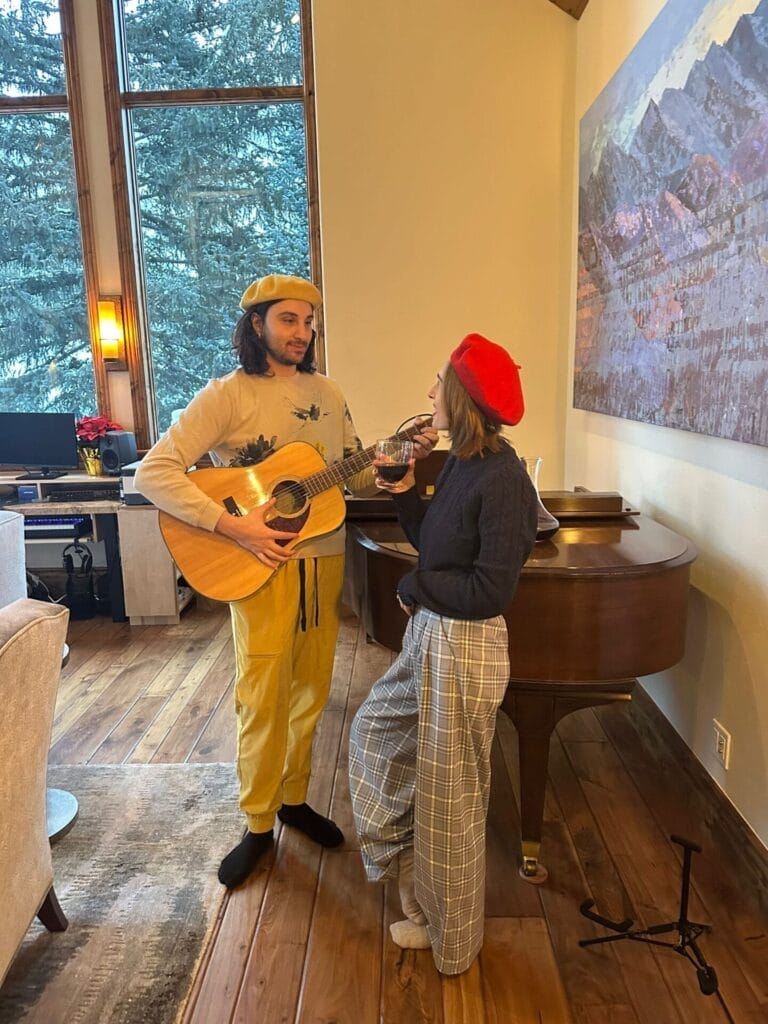 Tariq Abou-Bakr playing the guitar while standing in front o a piano in a room with a hard-wood floor, with his partner Jeanee Crane-Mauzy standing before him, as if he's serenading her.