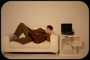 Press photo of Rob Eberle, wearing a brown outfit, lying on a white couch, with a record player to his left, that's on a white table.