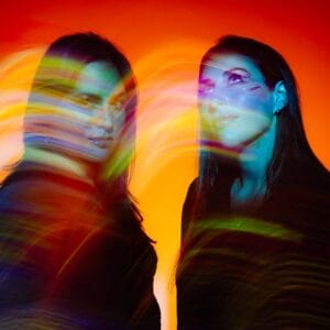 Official single cover artwork for "When Does Heartbreak Go Home" which shows the duo Royal She posing in front of an orange background while orange and blue light-effect filters are displayed in the foreground.