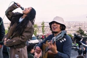 INNA holding the microphone in the air, leaning backwards and singing, with Sergio Muñoz on the right, sitting down playing his guitar, while wearing a white bucket hat. They're performing on the steps of Sacré-Cœur.