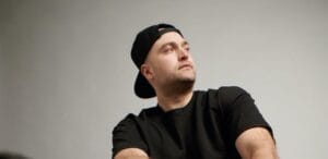 Photo of Seve Clain wearing a black cap on backwards, and a black t-shirt, with a grey background behind him.