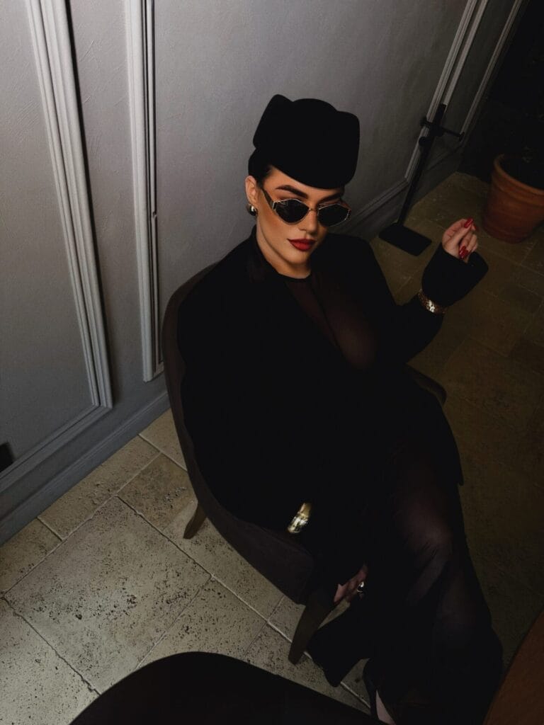 The Future of Fashion: Ukrainian Blogger Galina Denisiuk on Myths, Affordable Style, and the Art of Dressing Well 50 Galina Denisiuk sitting on a chair and looking up at the camera and she is wearing a black jacket over a black tight mesh dress with black heels along with a black hat and shades.