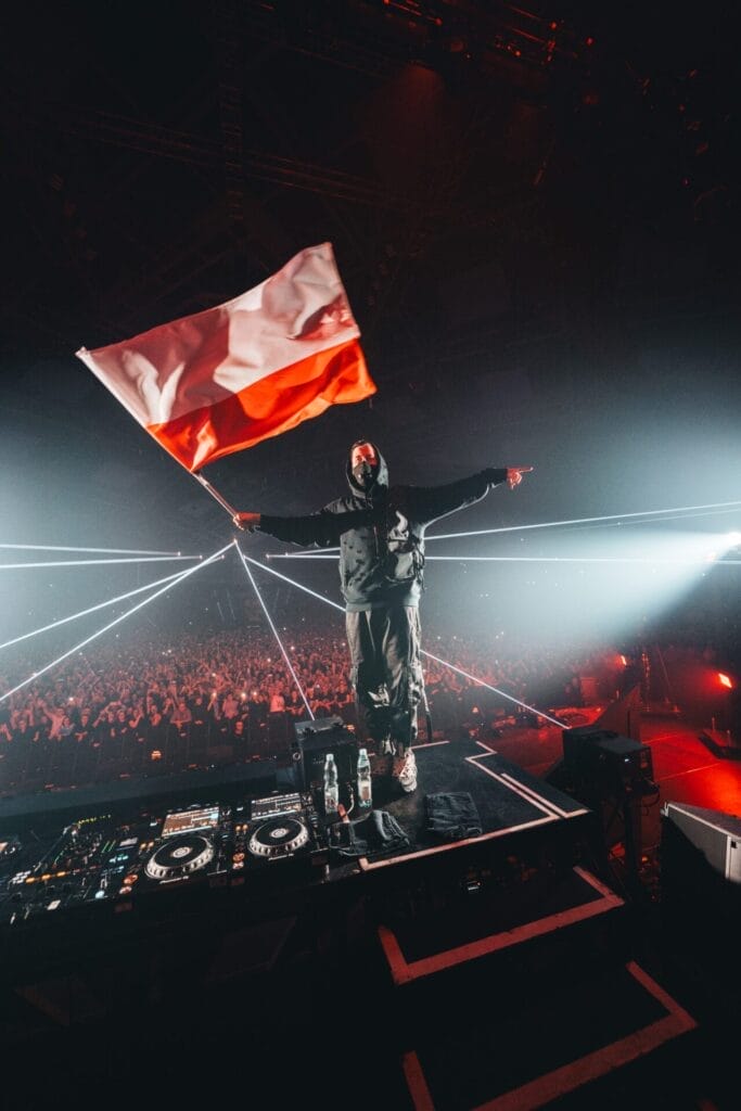 Alan Walker at Walkerworld in Łódź, Poland, standing on the DJ deck stage whilst holding a giant Polish flag in the air.