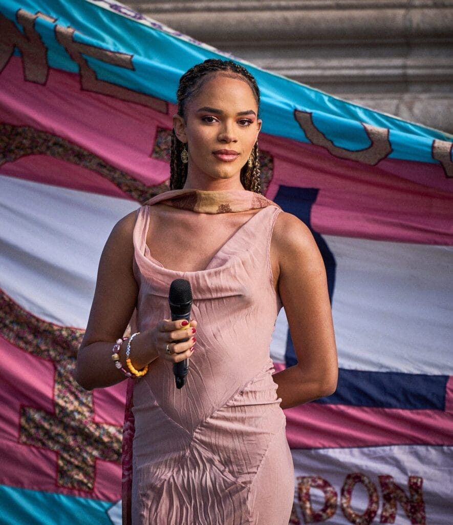 Yasmin Finney with a microphone in hand speaking at the London Trans+ Pride 2024. There's a huge trans flag behind her.