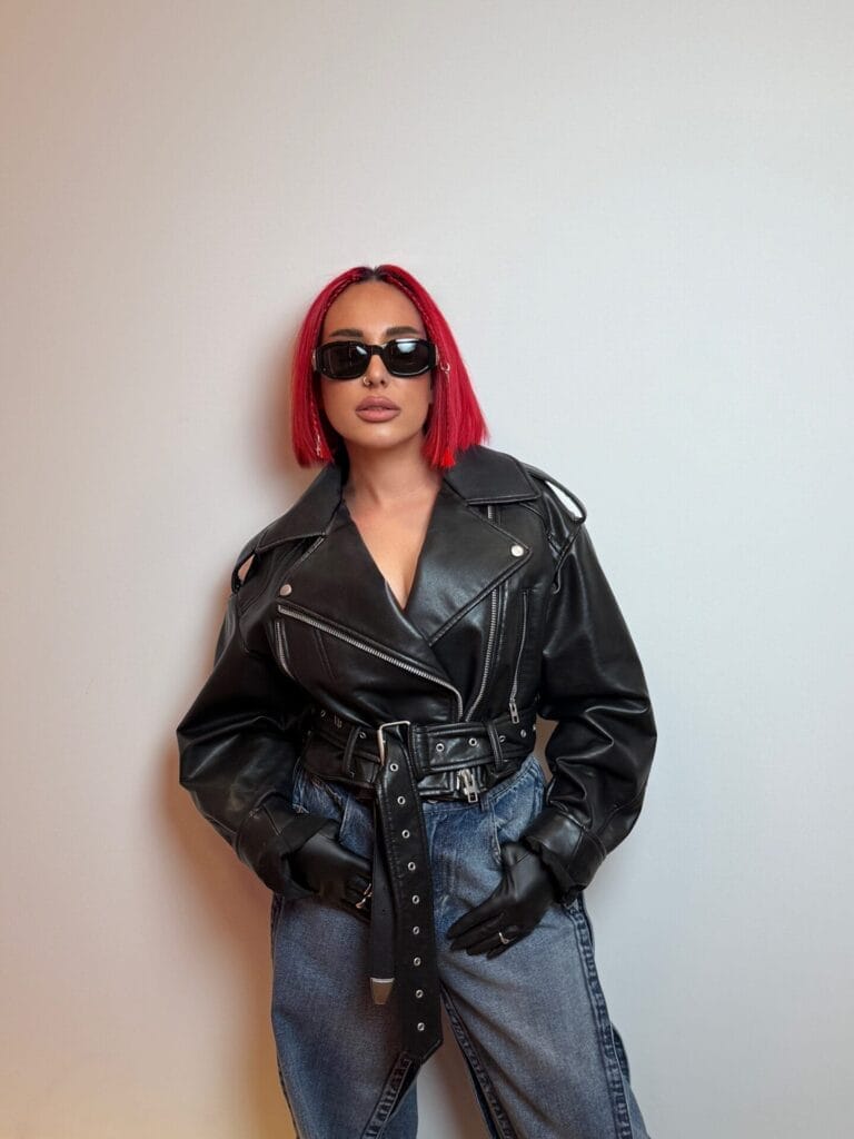 Interview: SHADU is the Ukrainian DJ Who is Redefining Afro House With Global Rhythms and Studio Soul in Hit Single "Contigo" 28 Press photo of SHADU who still has her red hair styled in a bob and she still has her shades on, but this time she's wearing a black leather biker jacket with black gloves and a pair of blue jeans.