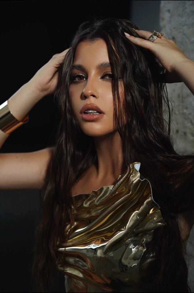 DaRina Ulanovska Captures the Fragility of Real Love with New Single “Лілеї" Meaning “Lilies” 7 Promotional photo which sees DaRina Ulanovska wearing a gold-metallic top with a matching bangle on her wrist, while her hair flows wet down the sides of her face. She has her hands posed on the top of her head.