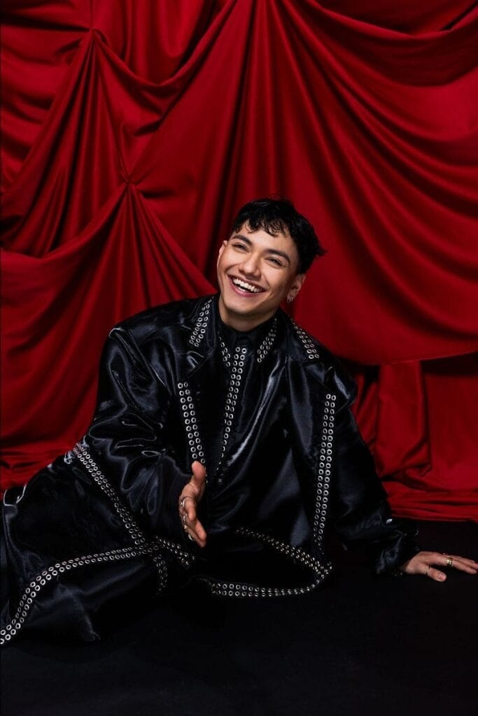 Original full portrait image of Eurovision 2025 winner JJ wearing a black outfit, sitting on the floor and resting on his left hand, while holding out his right hand to shake hands, while smiling. Behind him is a red curtain bunched up to look royal.
