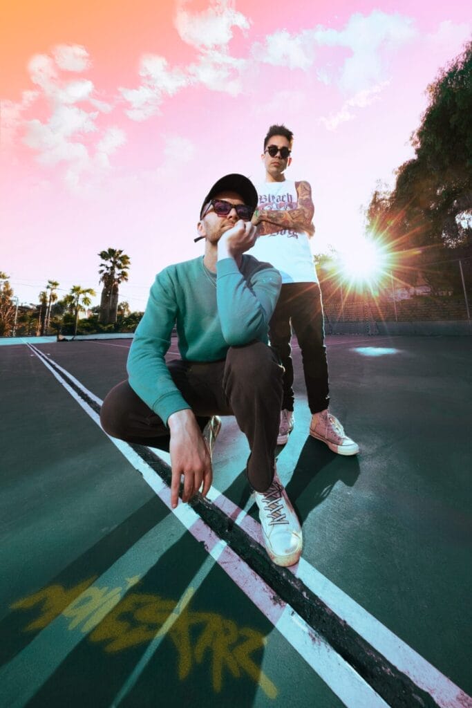Full portrait image of the official press photo of MOVIESTARZ posing in a car park with a sunset sky behind them. One of the guys are crouched while the other is standing behind him with arms crossed.