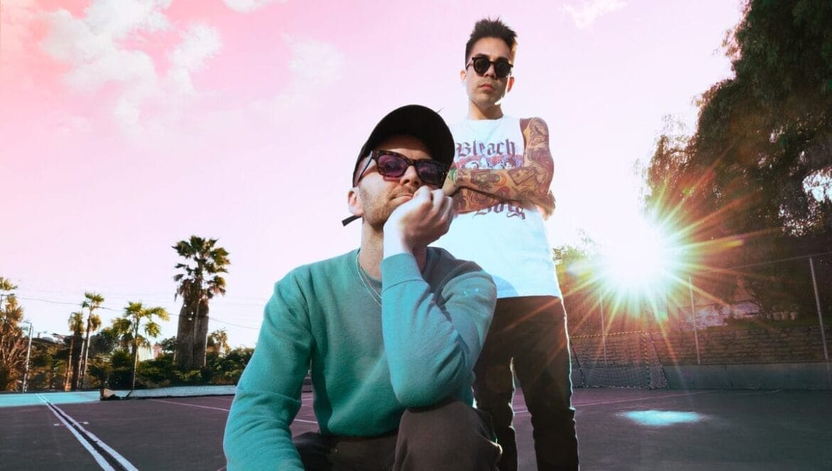 Official press photo of MOVIESTARZ posing in a car park with a sunset sky behind them. One of the guys are crouched while the other is standing behind him with arms crossed.