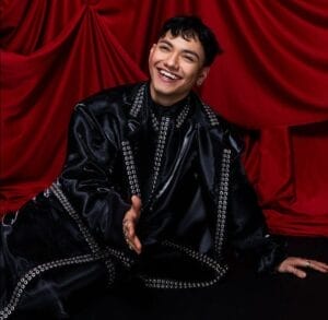 Cropped image. JJ wearing a black outfit, sitting on the floor and resting on his left hand, while holding out his right hand to shake hands, while smiling. Behind him is a red curtain bunched up to look royal.
