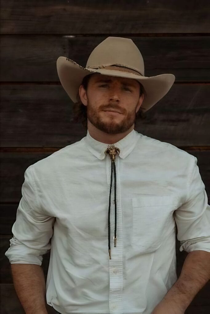 Full portrait version of the press photo for the announcement of the Grand Ole Opry. Callum Kerr in a white shirt with a golden bolo tie which is in the shape of a ram. He's also wearing a beige cowboy hat and is standing against a wood panel.