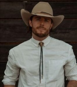 Cropped version of Callum Kerr in a white shirt with a golden bolo tie which is in the shape of a ram. He's also wearing a beige cowboy hat and is standing against a wood panel.
