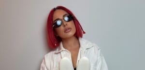 Head shot photo of SHADU who has red hair cut in a bob. She is wearing a white jacket and sunglasses with a grey background behind her.