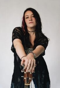 Press photo of Lindsay Jordan posing by standing behind her guitar, which is out in front of her, with her hands on the headstock of the guitar. She's dressed in black and there's a grey background behind her.