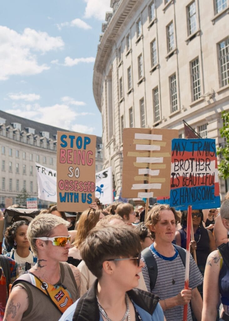 Crowd from London Trans+ Pride 2025 with signs that say "Stop being so obsessed with us" and "My trans brother is beautiful & handsome".