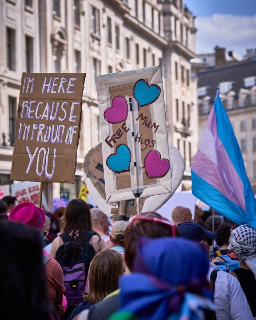 London Trans+ Pride 2025: Powerful Celebrity Messages Echo the Theme "Existence & Resistance" 111 A crowd photo from London Trans+ Pride 2024 which includes a trans flag in the air along with two signs, one that says "I'm here because I'm proud of you" and another that says "Free Mum Hugs".