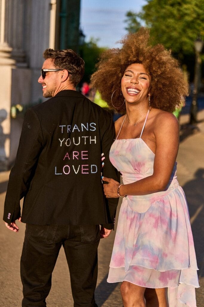 Jude Guaitamacchi and Olivia Campbell-Cavendish posing together at London Trans+ Pride 2024 with Jude wearing a suit with the jacket having the words "Trans youth are loved" embroidered on the back, and Olivia has linked arms with him and is smiling while wearing a dress in a tie-dye colouring of the trans pride colours.