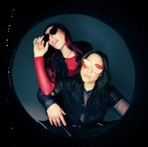The duo, Royal She, posing together, one behind the other, wearing heart-shaped glasses. There's a circular frame around the image.