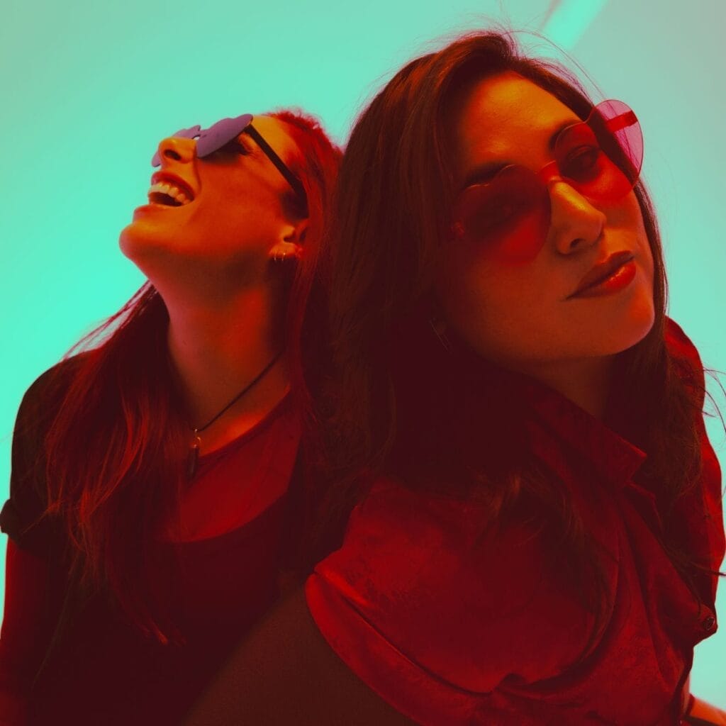 Official single cover artwork for "Feel Again", which sees Royal She posing in front of one another with a turquoise background and a filter that over-exposes the duo with an orange tone. They are both wearing heart-shaped glasses.