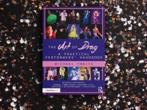 The book "The Art of Drag: A Practical Performers' Handbook" by Michael Twaits which is a purple cover with photos of drag queens, which is laying on a background of silver and black sequins.