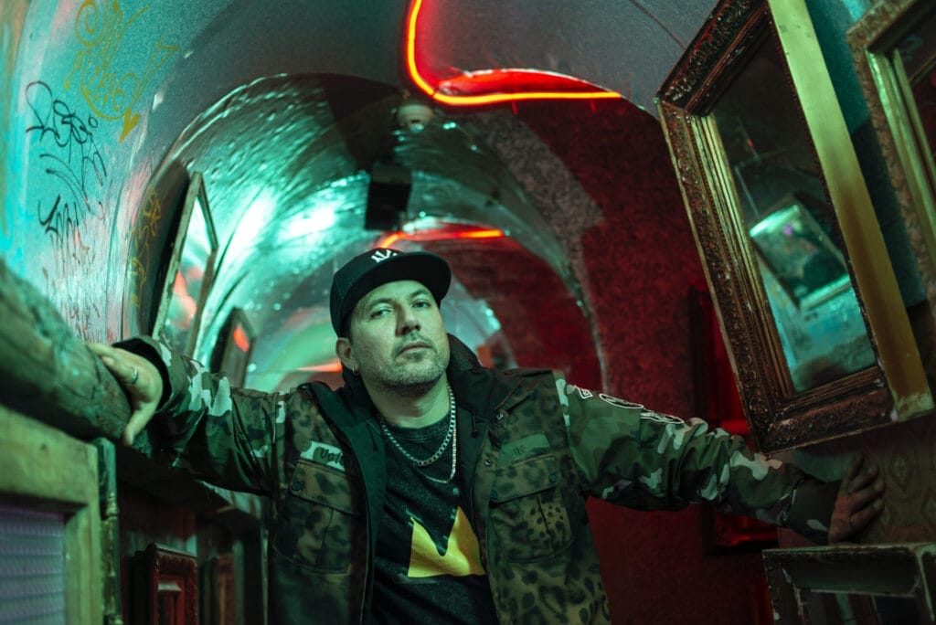 Victor Vildósola, who leads THEKOIWAY, in a small hallway that has a dark-lit feel, with mirrors on the right and graffiti on the right, and he has his arms out touching both walls, wearing a camo jacket and a black cap.