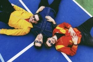 The trio, Daniel Ellsworth & The Great Lakes, lying on a blue court, with each of them wearing different colour jackets—yellow, blue, and red, along with shades.