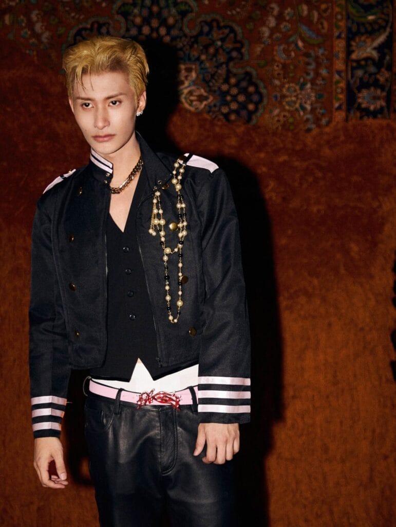 BENZO with blonde hair, looking smouldering into the camera as he wears a black bomber jacket over a black waistcoat over a white shirt, with some black trousers paired with a white belt.