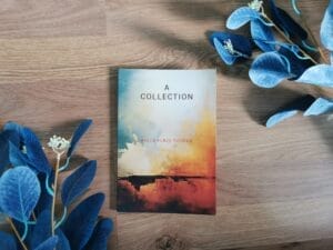 The book, "A Collection" by Becca Furze Thomas, lying on a wood background cornered by blue leaf plants.