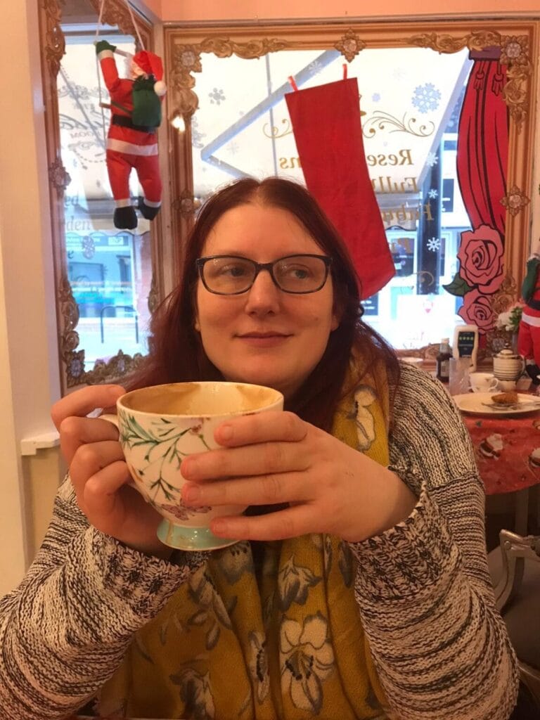 Book Review "A Collection" by Becca Furze Thomas: A Soulful Journey Through Life in Verse 4 Photo of Becca Furze Thomas in a café, holding a huge mug in her hands and wearing a jumper. There's a Santa climbing the window behind her, so it's around Christmas time.
