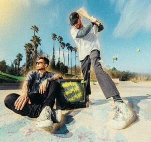 The duo, MOVIESTARZ, on the beach with palm trees in the background. One of the guys is sitting on the sand with an arm on a TV that has graffiti on, and the other member of the duo is standing up who is about to swing a baseball bat down over his head.