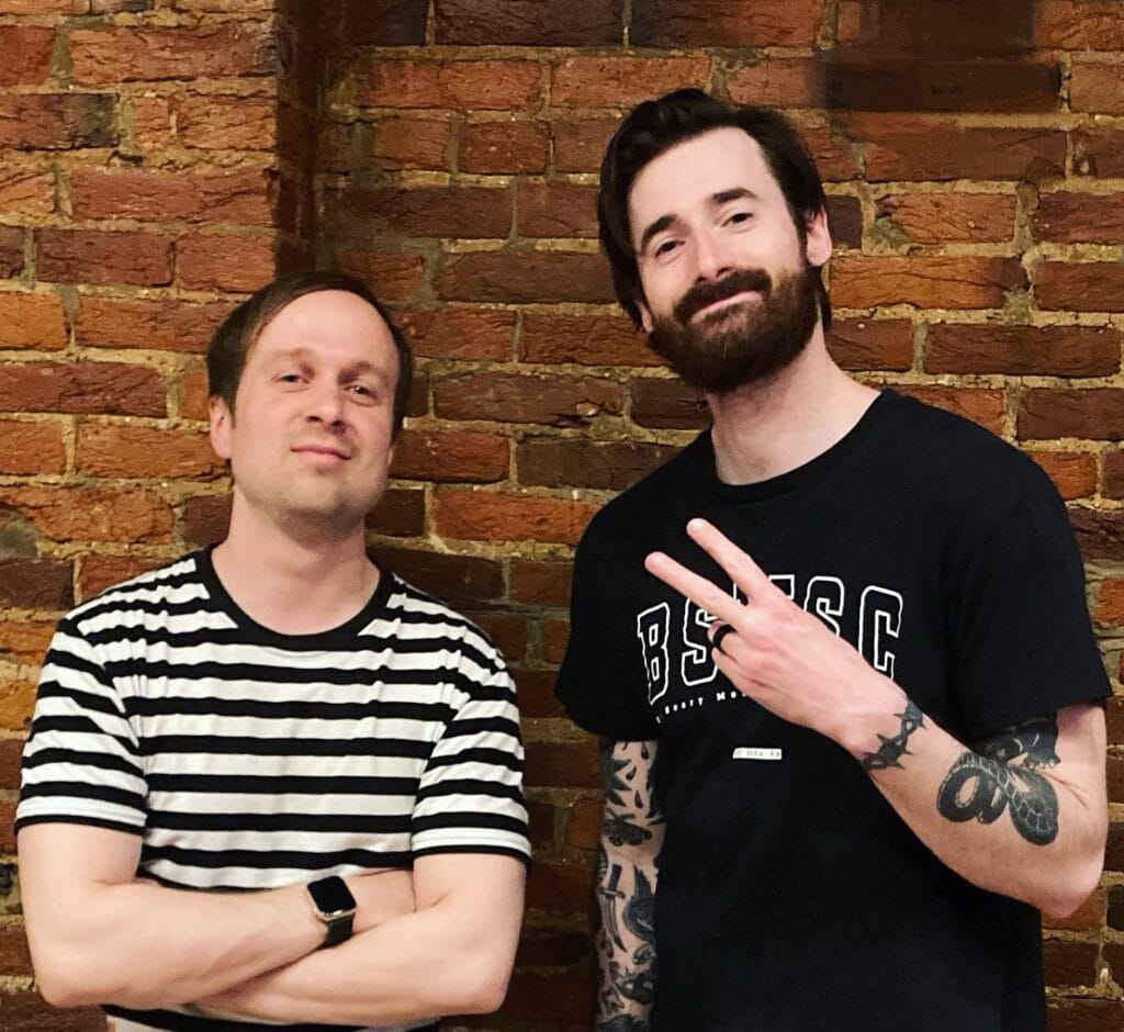 Official press photo of Joe Stanton and Mitchell Layton posing in front of a brick wall with Stanton wearing a black and white striped t-shirt with his arms crossed and Layton wearing a black t-shirt with his hand out in a peace sign.
