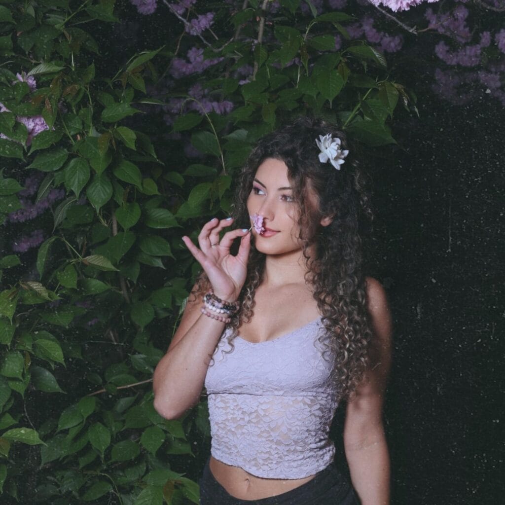 Nikki Silva Steps Into the Spotlight With Bold and Honest Debut Album "ty, places" 2 Official album cover artwork for "ty, places" which sees Nikki Silva posing in front of a bush, wearing a white vest-like top and a white flower in her hair, while holding a flower up to her nose.