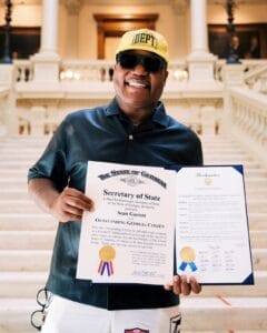 Sean Garrett smiling outside an official building's steps, holding the two state awards he received, while wearing a dark green shirt, a yellow Gallery Dept. snapback cap, and some shades.