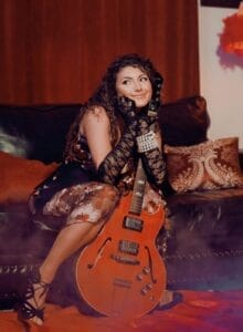 Nikki Silva posing with a guitar while sitting on a couch.