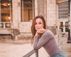 Meg Whalen wearing a grey top and jeans, resting her elbow on a metal bar with her hand under her chin as her auburn hair falls down her other shoulder. She looks like she's in a courtyard with a whit stone building behind her.