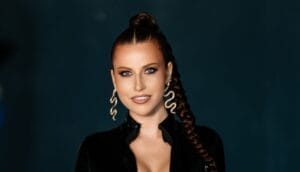 Cropped photo of a headshot of ZHANNET who is wearing a black outfit with her hair tied back and flowing down one shoulder in a plait, and she is wearing some big gold snake earrings.
