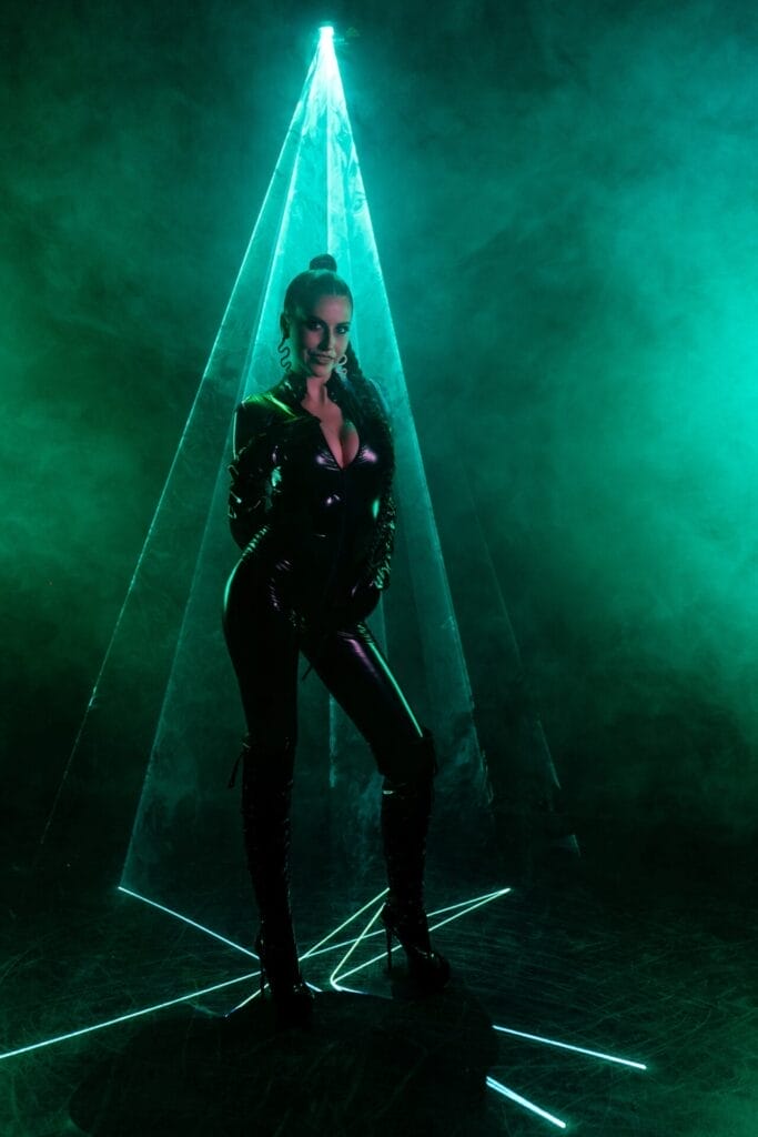 ZHANNET wearing a black latex bodysuit standing in the middle of a light green laser light that is facing down on her with a smoky atmosphere creating green smoke as the laser bounces around the room.