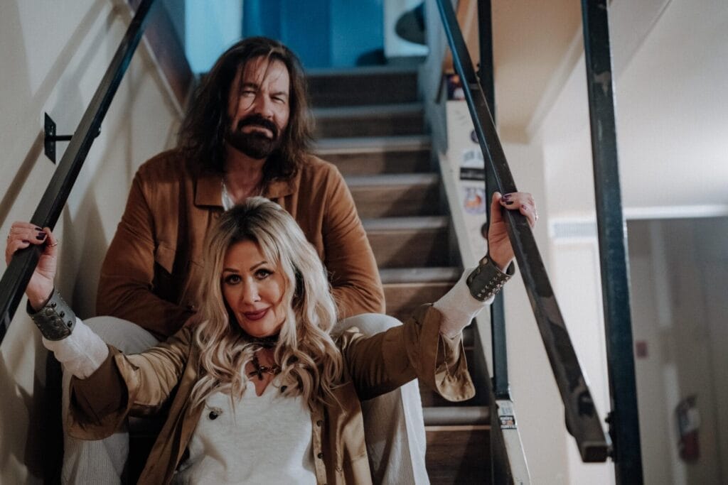 The founding members of D2UR, husband and wife Diane and Mike Isbister, posing on a set of stairs with Diane sitting in front of Mike, in the backstage area of a venue.