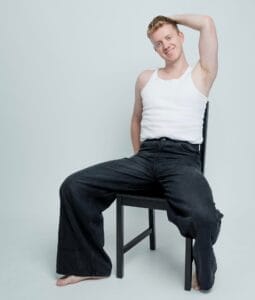 James Barr posing for a professional photo shoot, sitting on a black chair, wearing a white vest top and black flare jeans. He has one hand behind his head