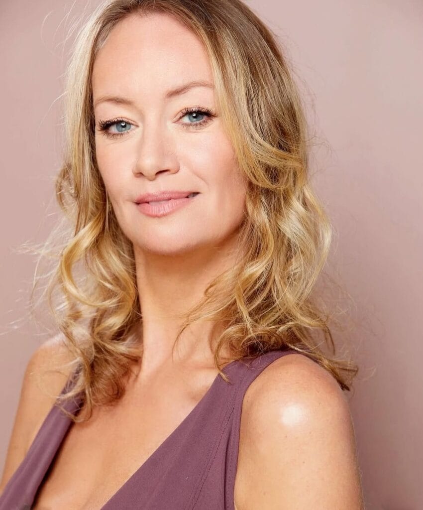 Head shot of Joanna Pickering who is smiling at the camera as her blonde hair curls down past her shoulders. She's wearing what looks to be a purple dress or top.