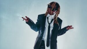 Lil Wayne posing for a professional photo shoot wearing a white shirt that is outlined along the collar, a black tie which hangs loosely around his neck, and a black suit jacket along with some shades. He is smoking and has his hands up in a shrug-like pose.