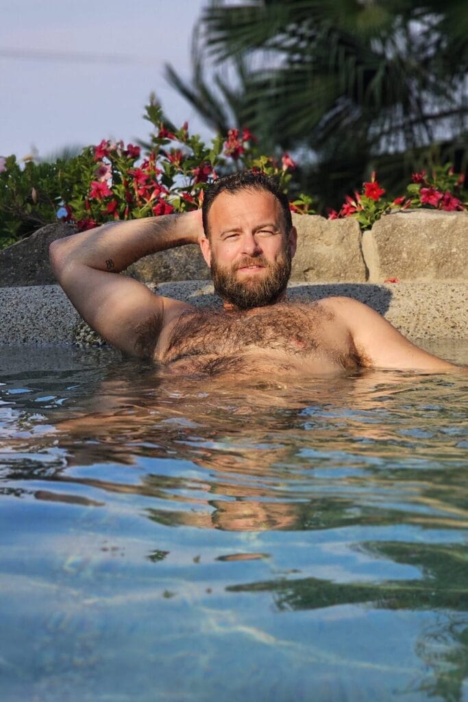 Mike Maimone relaxing in a pool with one hand behind his back, showing off his hairy chest.